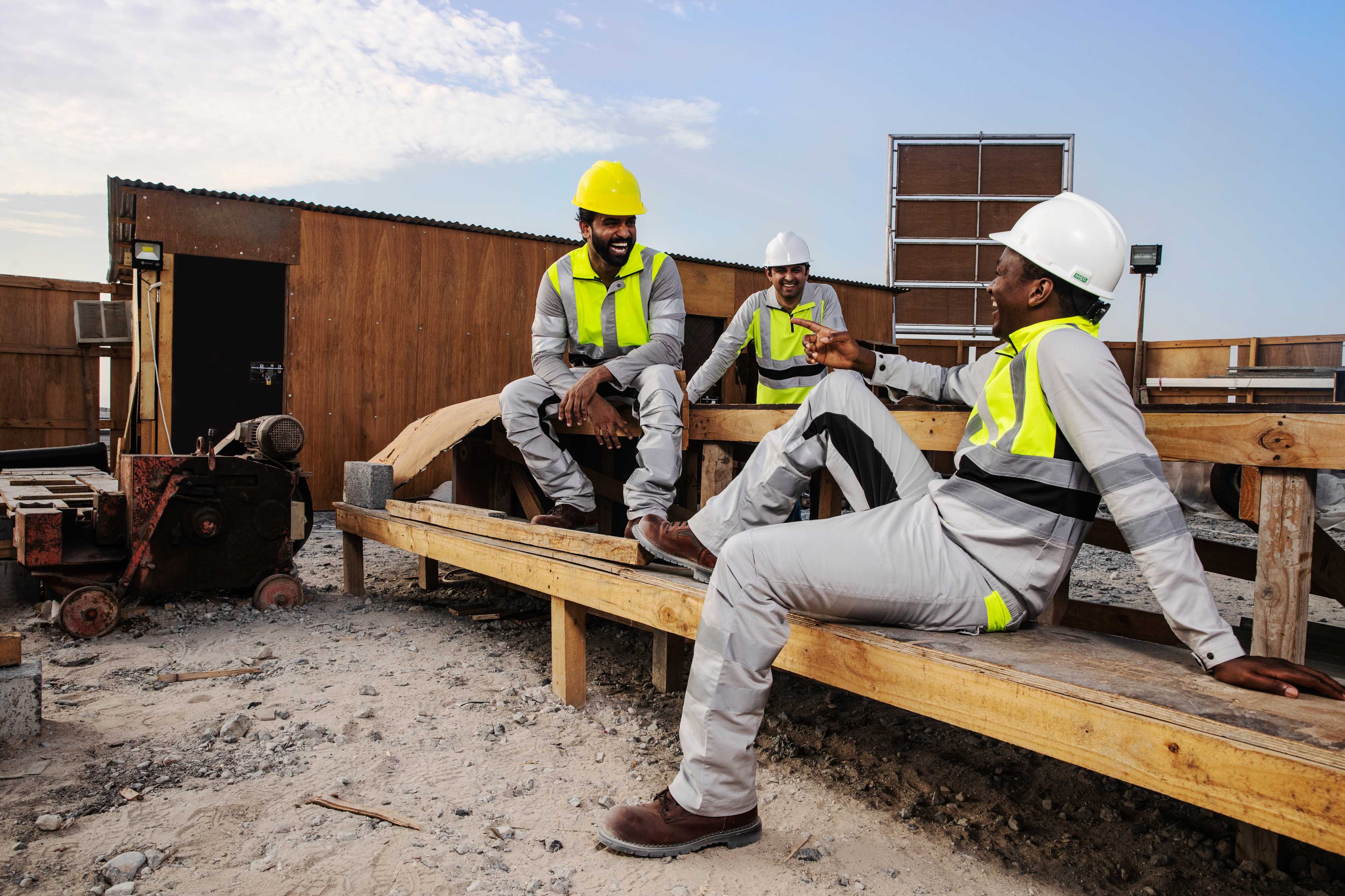 Construction crew in Shamal lightweight high-visibility workwear on a Middle East job site, showing reflective safety garments designed for extreme heat. Three construction workers wearing Shamal high-visibility workwear sit and talk on a job site during a break.